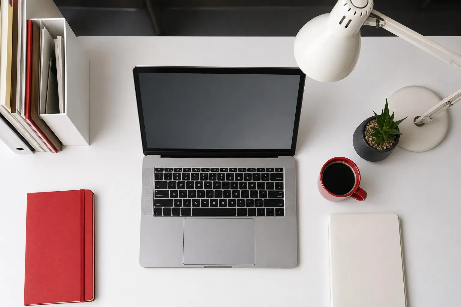 Laptop, desk lamp, potted plant, red notebook, coffee mug on a desk.