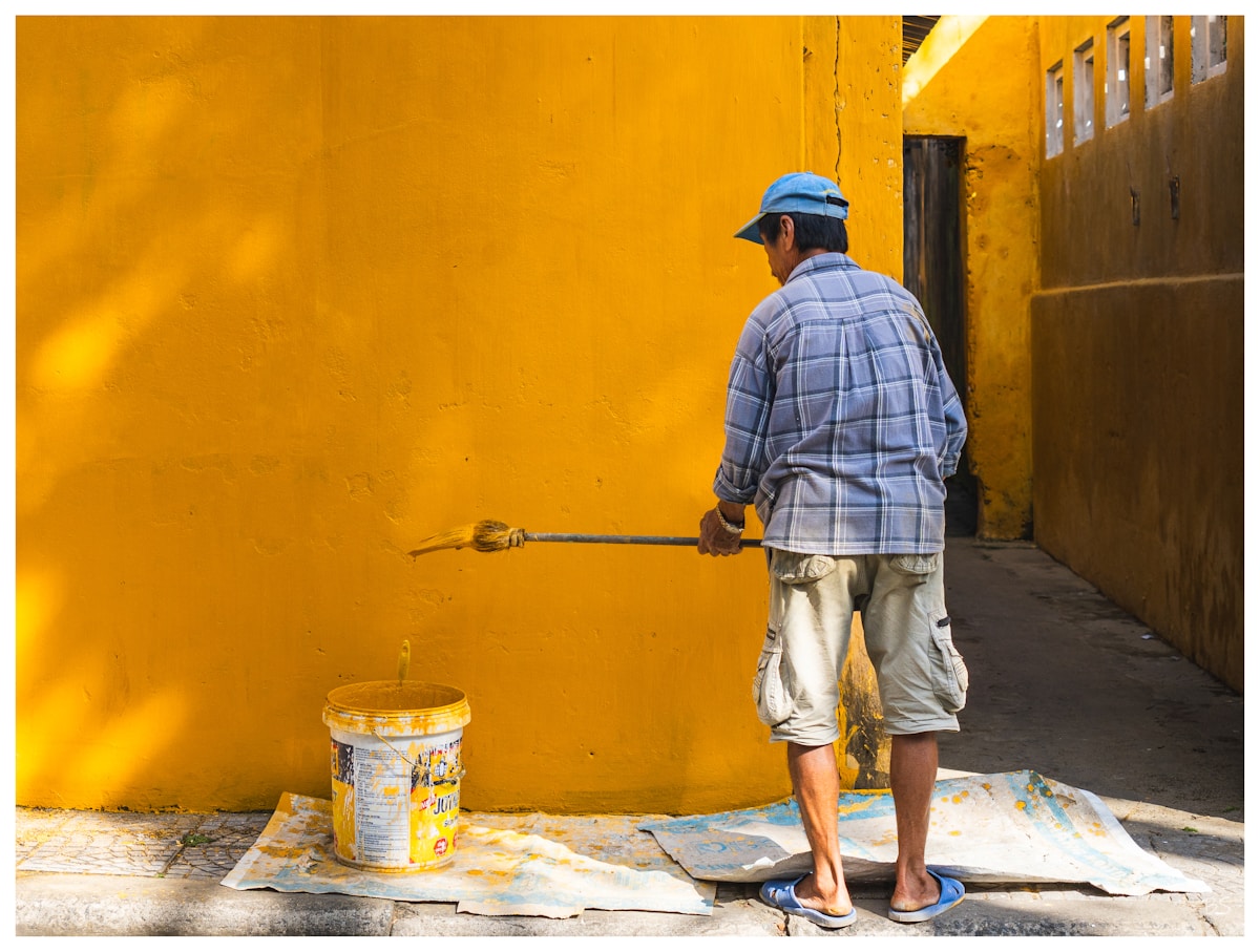 Homme peignant un mur jaune vif dans une ruelle étroite.