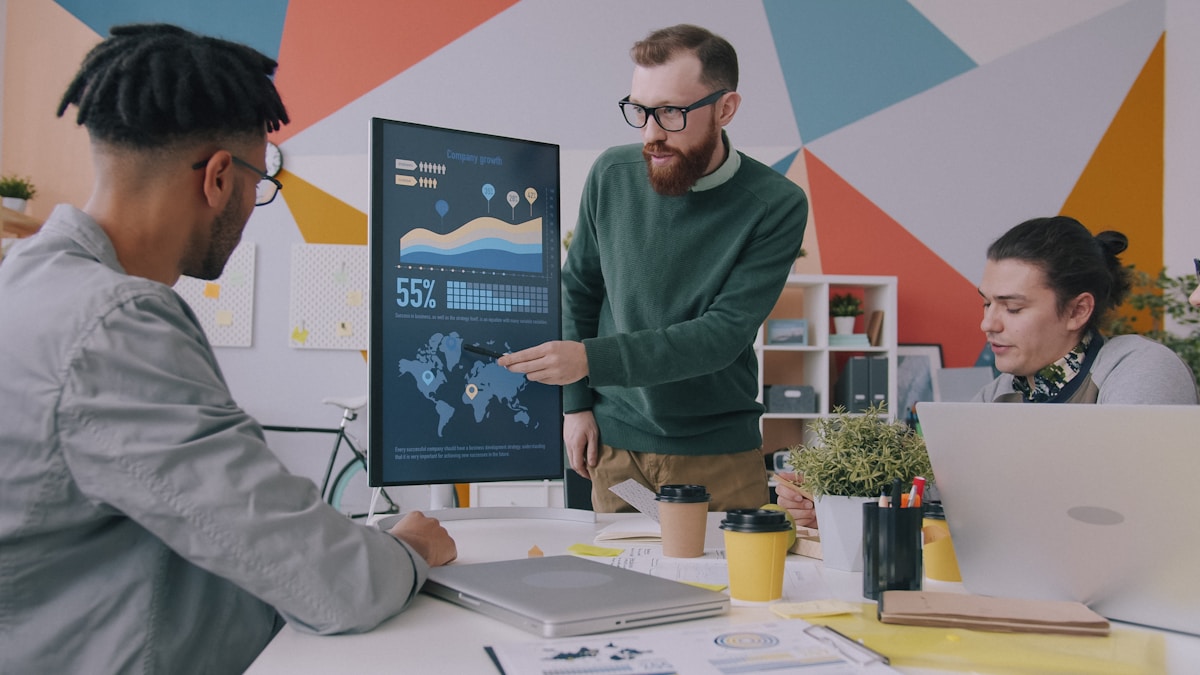 A man pointing at a vertical monitor displaying data charts to two colleagues