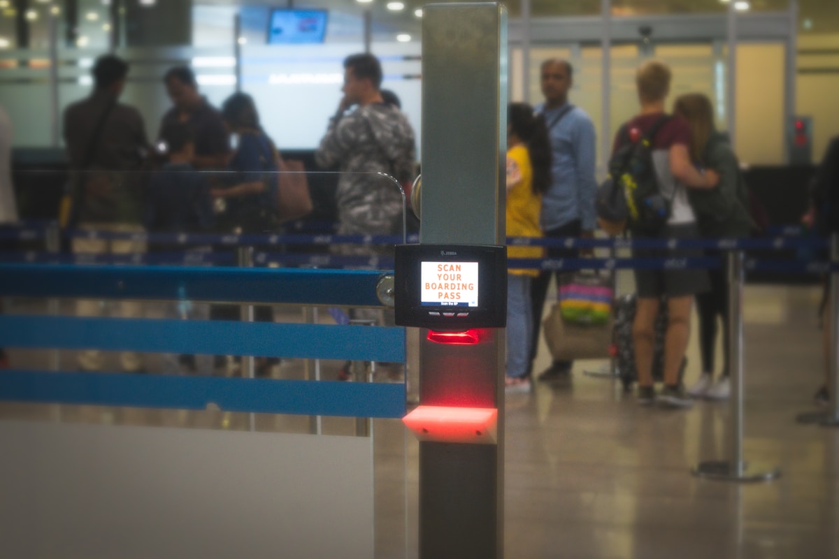 Borne de contrôle des cartes d'embarquement dans un aéroport avec des passagers en arrière-plan.