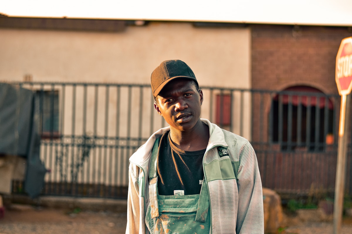 Un homme africain portant une casquette et un gilet rayé, debout dans une zone urbaine.