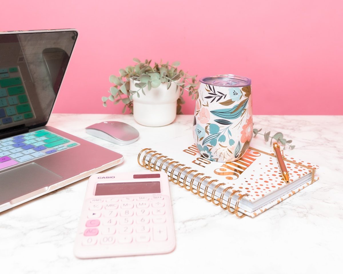 Bureau de travail rose et blanc avec ordinateur portable, calculatrice, cahier et tasse décorée.