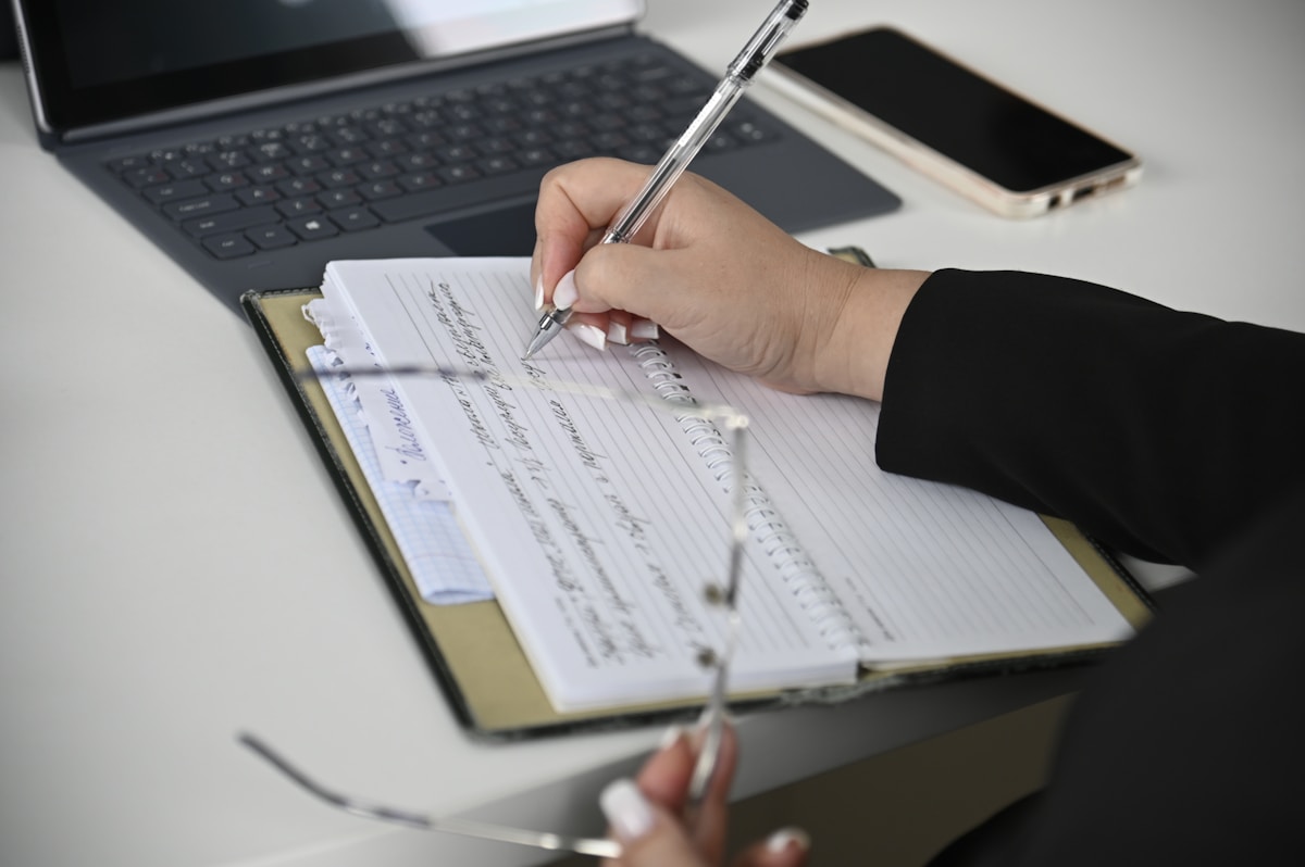 Personne écrivant dans un cahier sur un bureau avec un ordinateur portable.