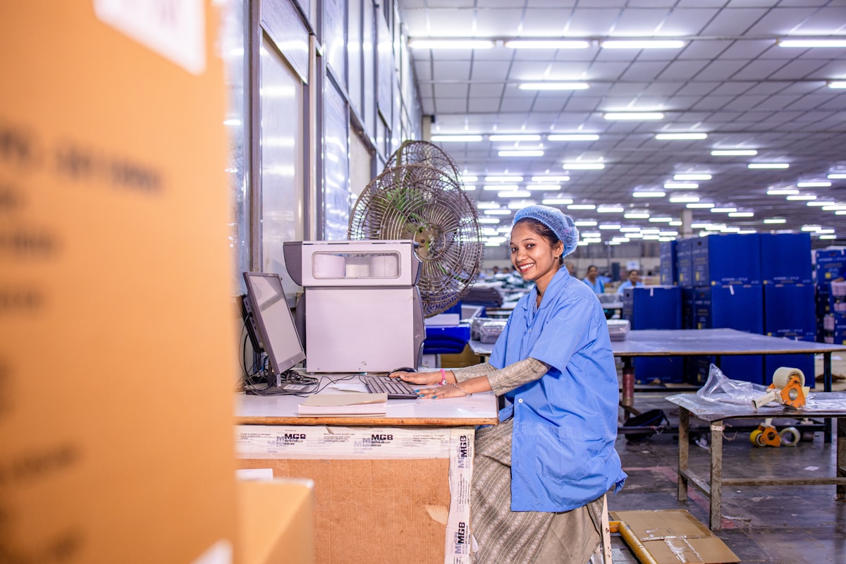 Employée souriante dans un entrepôt industriel, utilisant un ordinateur.