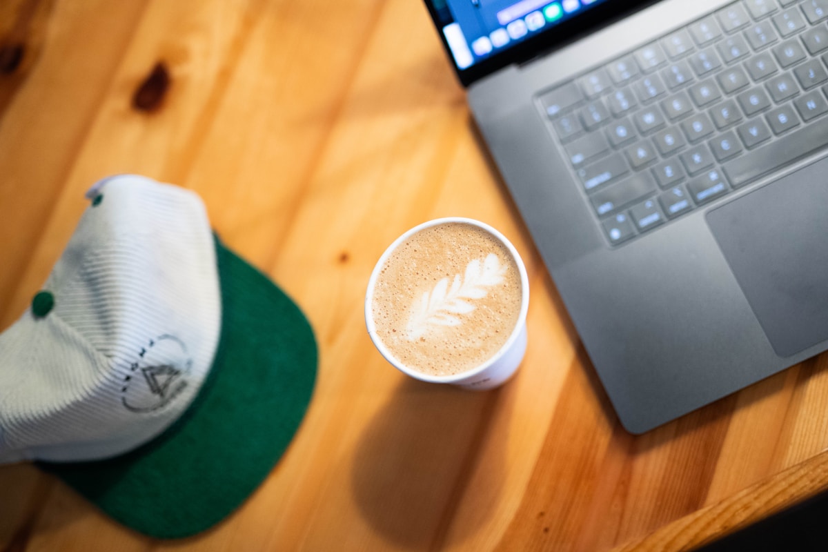 Tasse de café avec motif de feuille, ordinateur portable et casquette sur une table en bois.