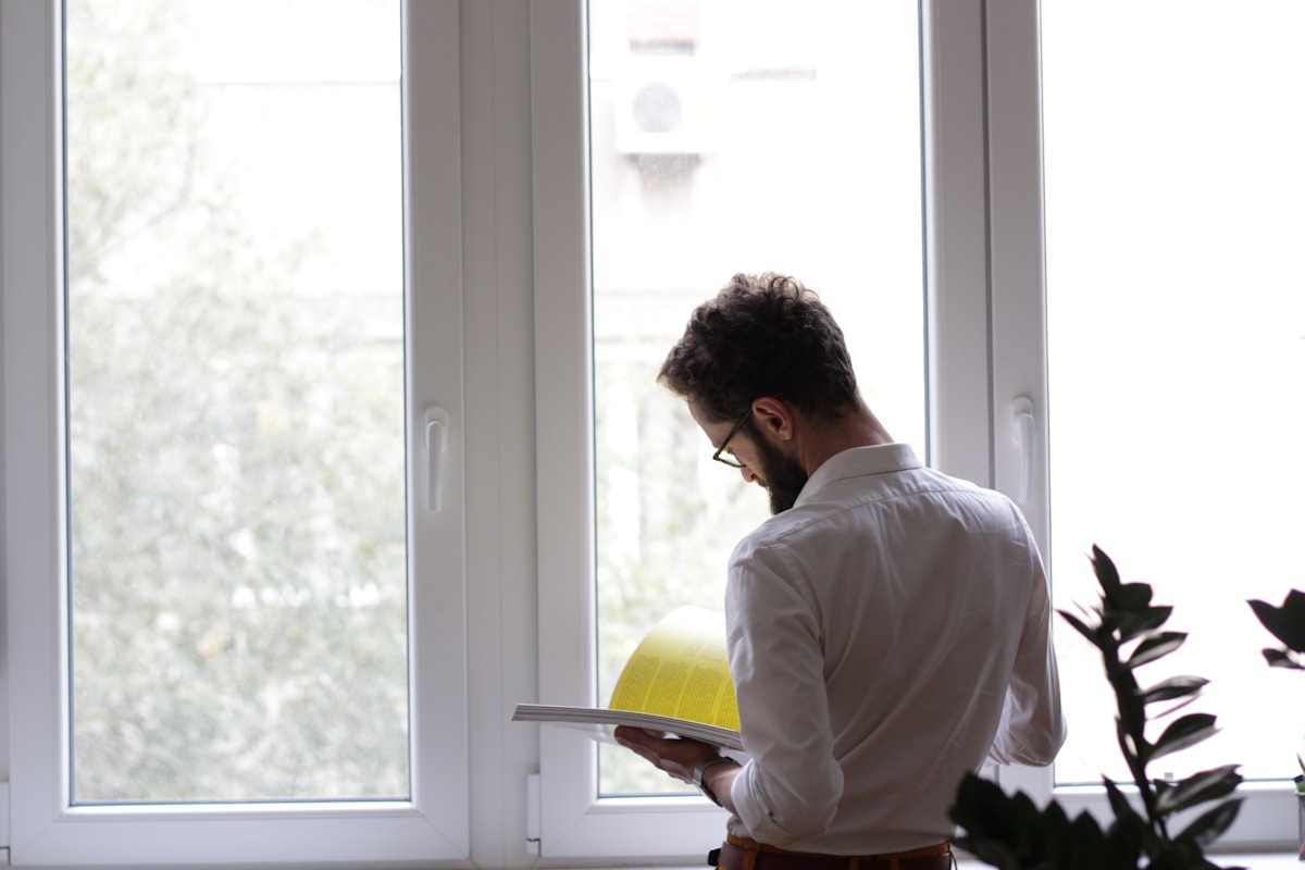 Homme en chemise blanche debout près de la fenêtre, lisant un livre.