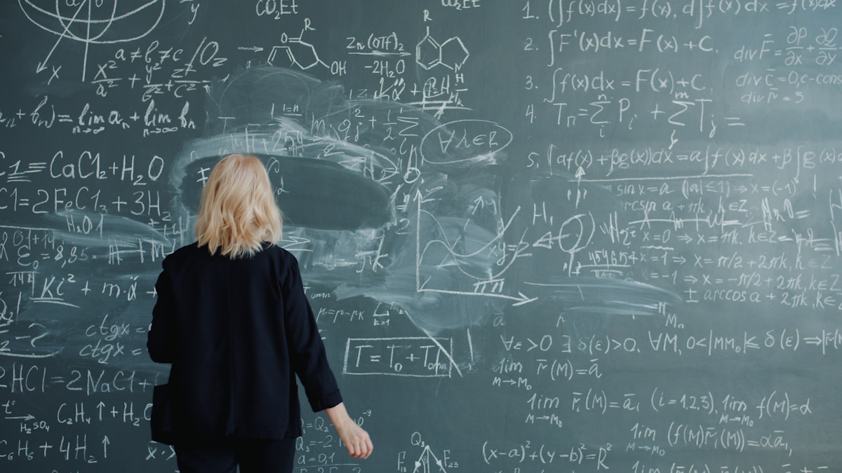 Femme blonde de dos devant un tableau noir rempli d'équations mathématiques.