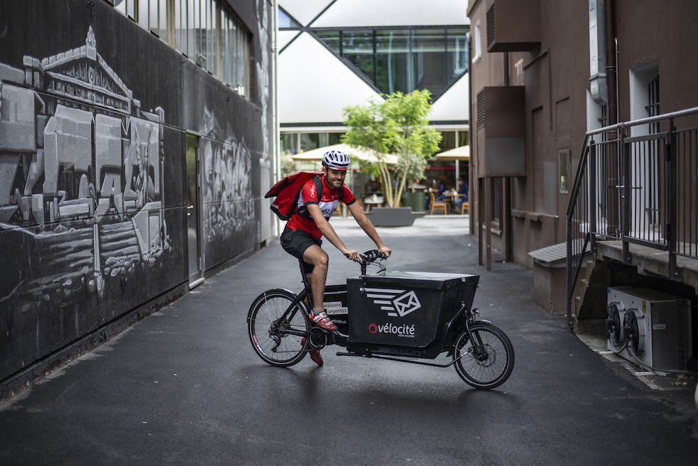Un cycliste avec un vélo cargo livrant des caisses de Grano Maté dans une rue de ville suisse.