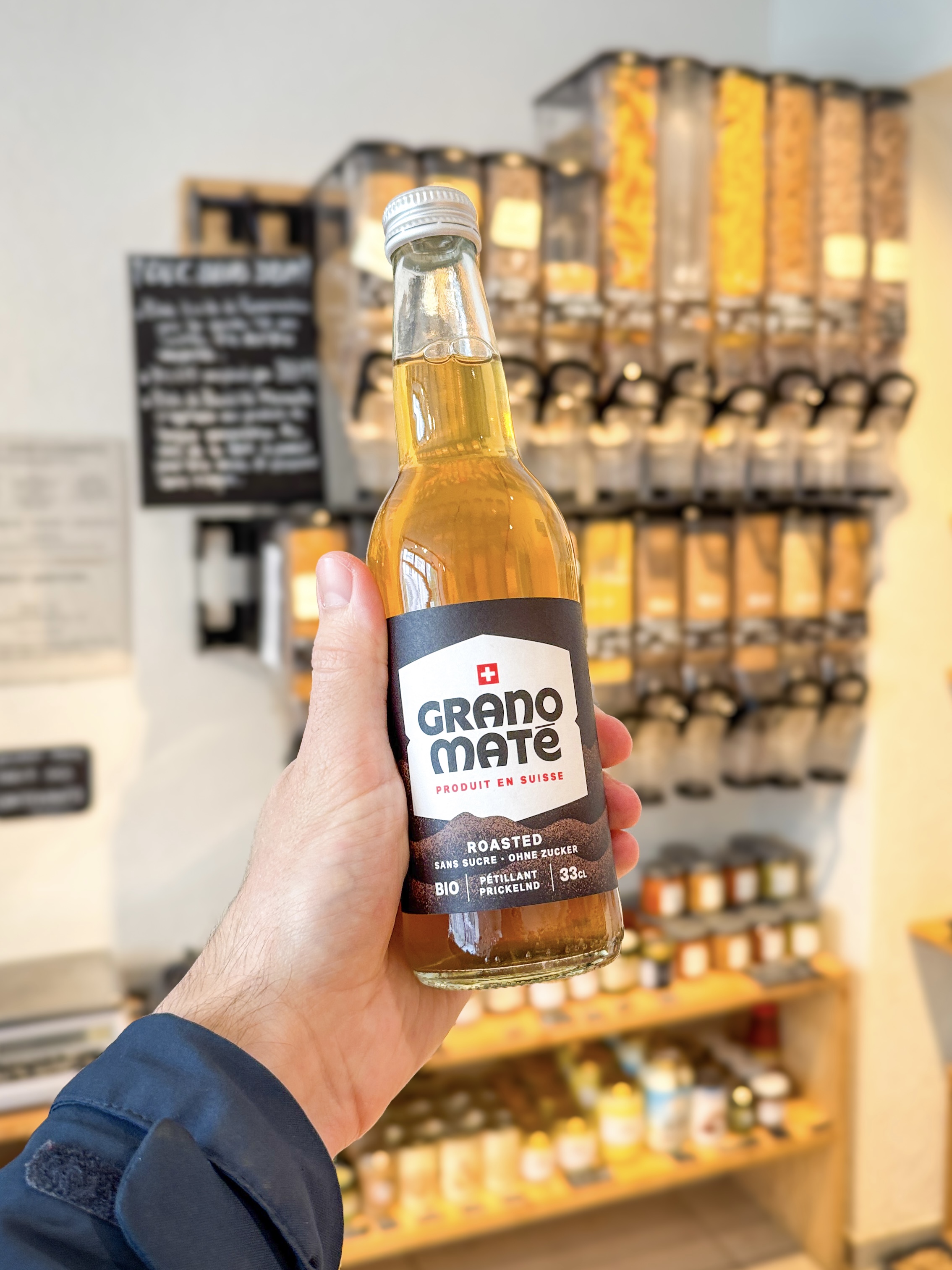 Une photo d'une bouteille de Grano Maté Roasted BIO et une canette de Grano Maté Gingembre posées sur une étagère en bois rustique dans une épicerie fine, avec un éclairage chaleureux.