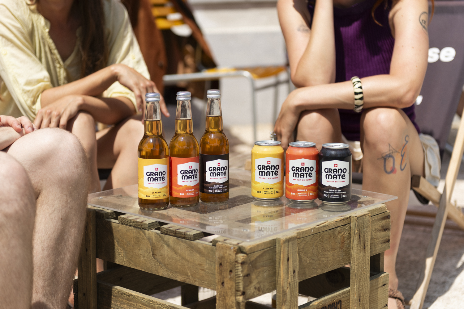 Image promotionnelle de Grano Maté. Plusieurs bouteilles et canettes des différentes saveurs sont disposées artistiquement sur une table en bois rustique. Trois personnes sont assises autour de la table.