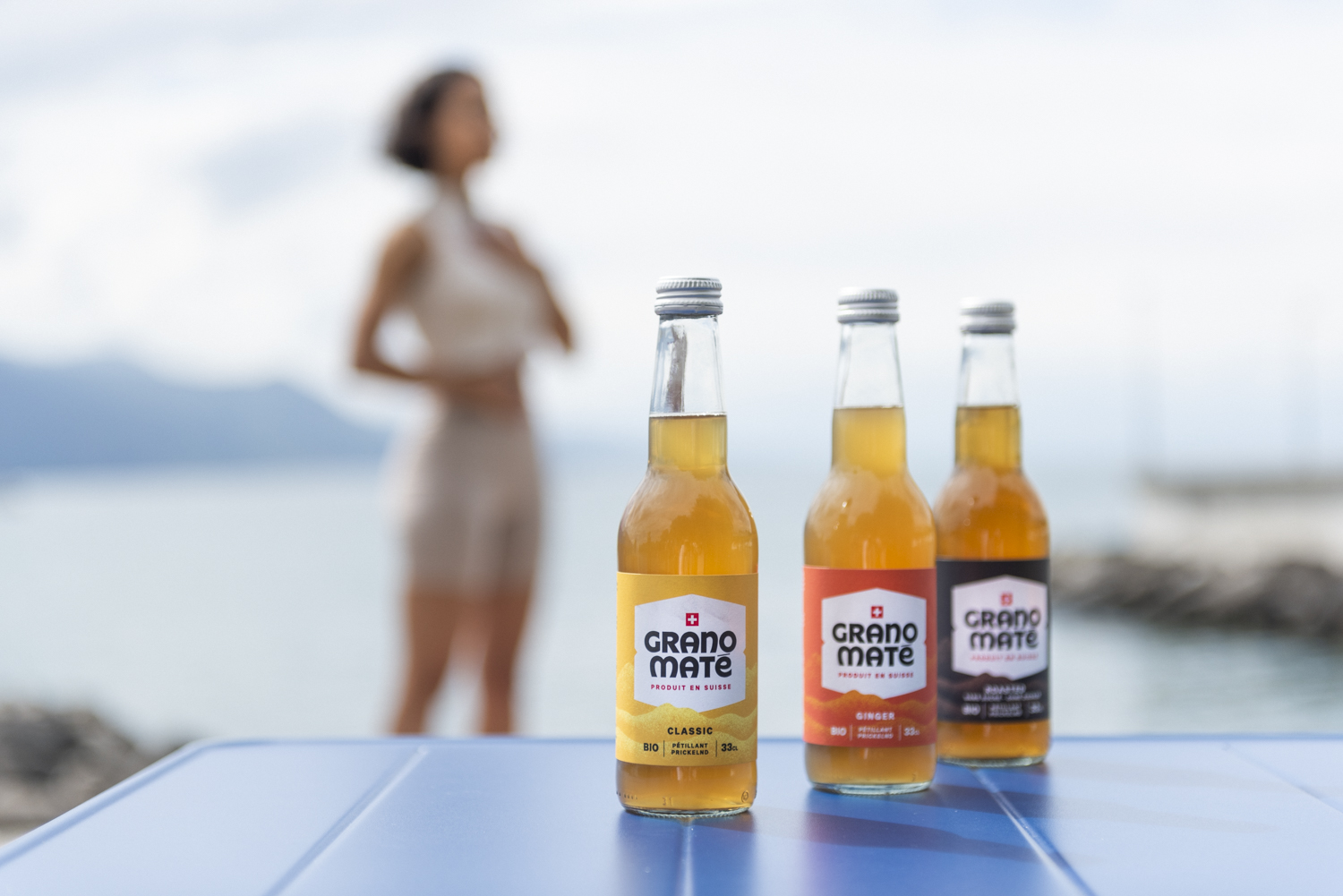 Trois bouteilles de Grano Maté au premier plan sur une table bleue, au bord d'un lac. Une silhouette féminine debout fait des exercices de bien être en arrière plan.