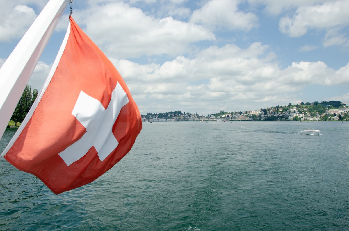 Drapeau suisse flottant sur un lac avec ville en arrière-plan.