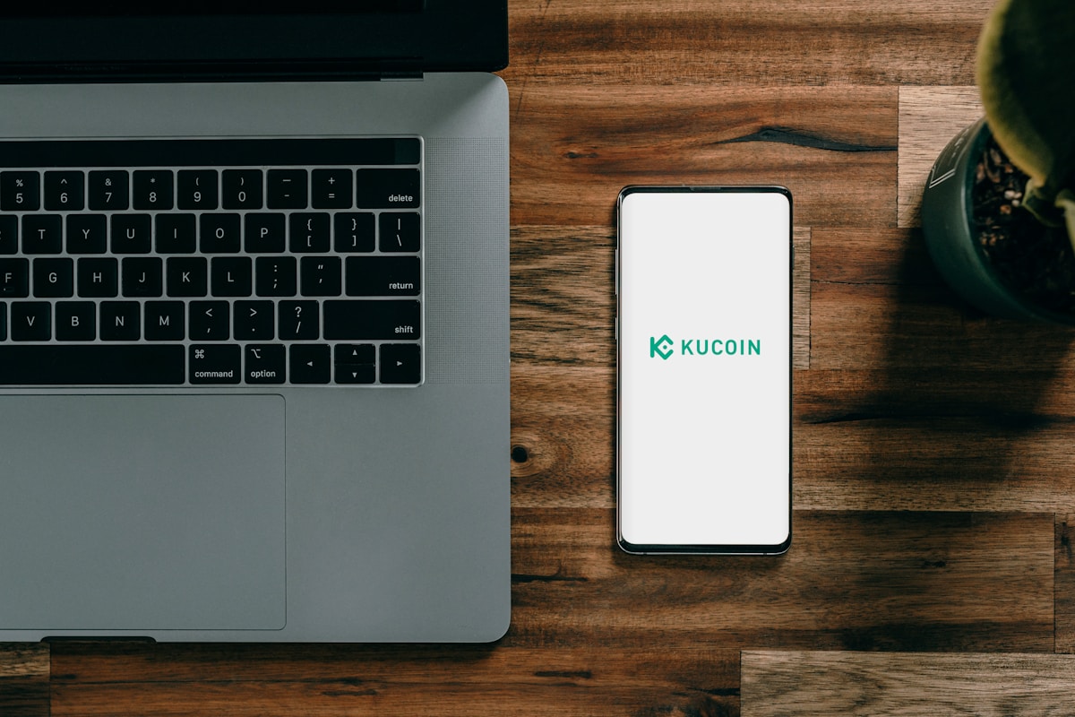 Ordinateur portable, smartphone avec logo Kucoin, plante verte, surface en bois.