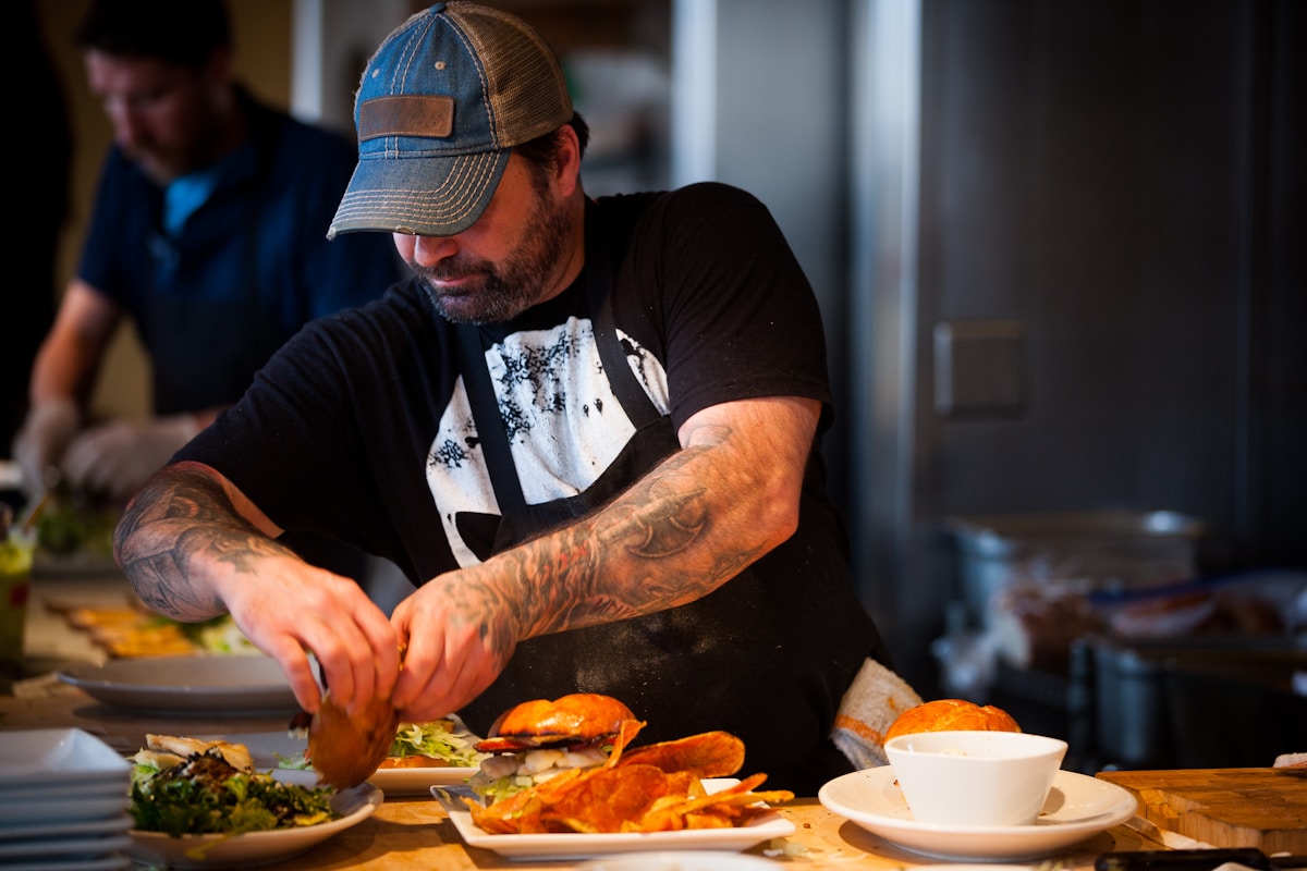 Homme tatoué préparant des hamburgers dans une cuisine.