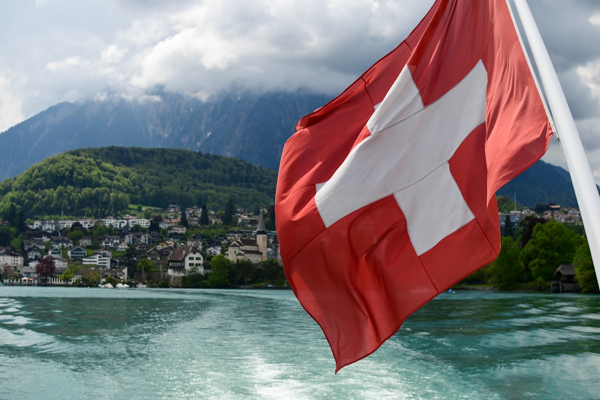 Drapeau suisse flottant sur un lac de montagne avec village en arrière-plan.