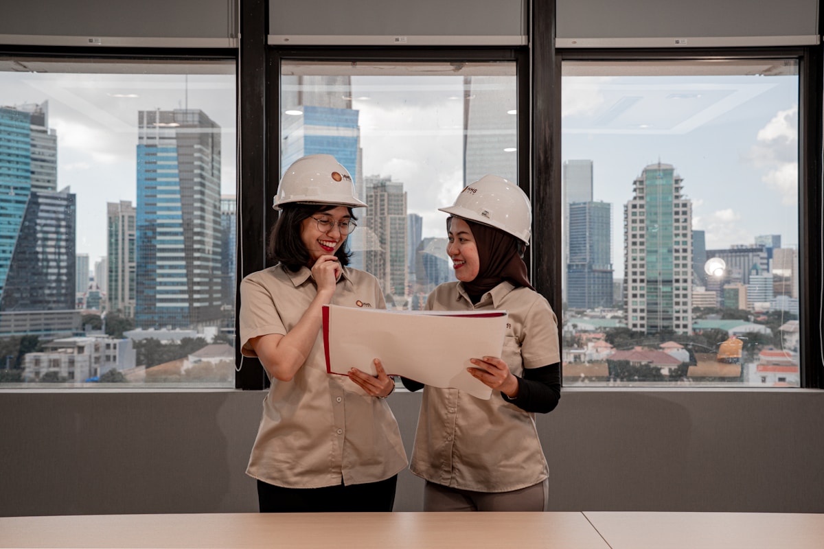 Deux femmes en casques de chantier examinant des plans dans un bureau avec vue sur la ville.