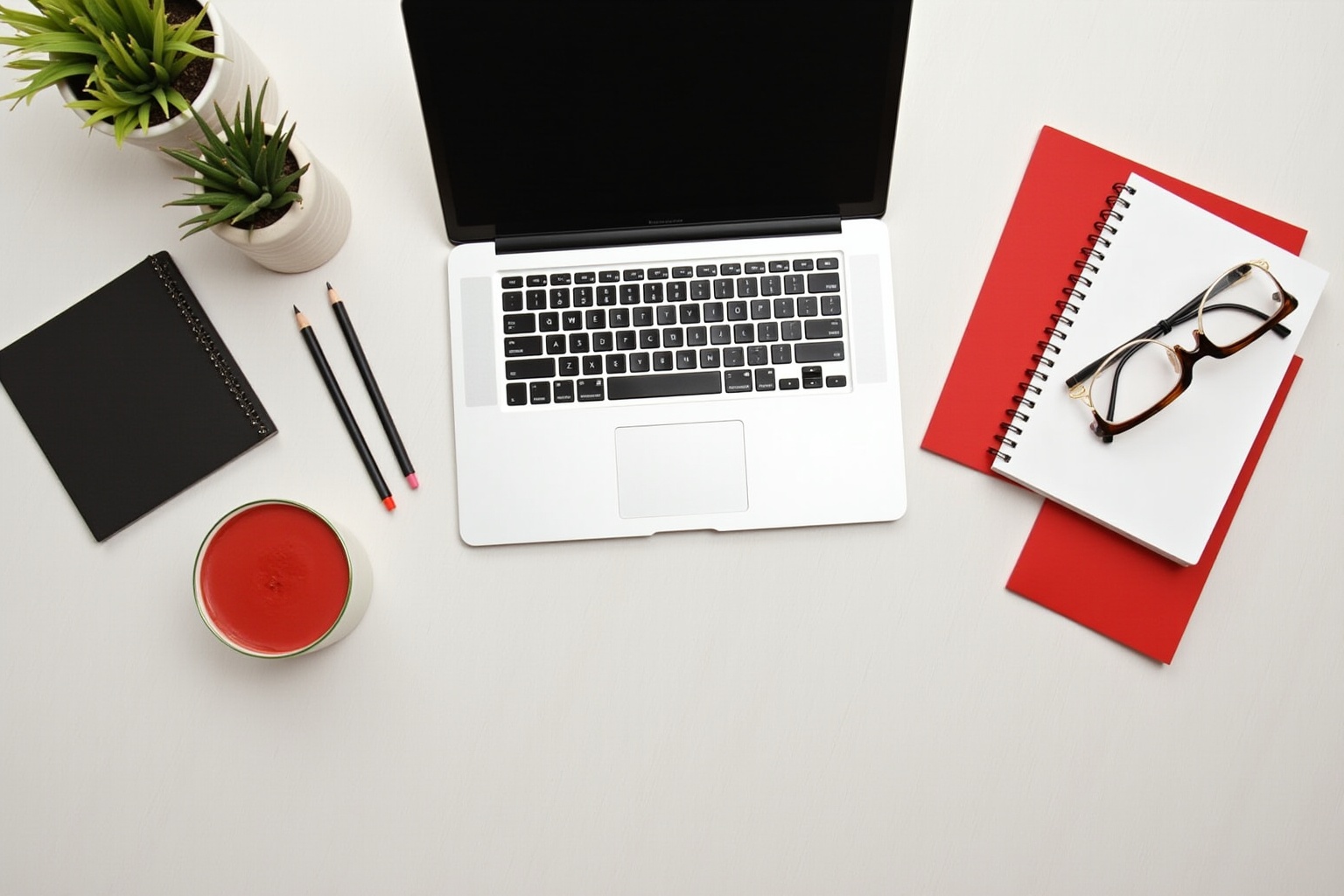 Laptop, notebook, pencils, plant, and glasses on a white desk.