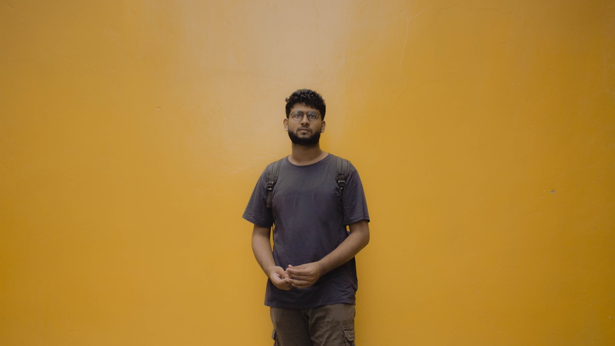 Homme debout devant un mur jaune, portant un t-shirt gris et un pantalon cargo.