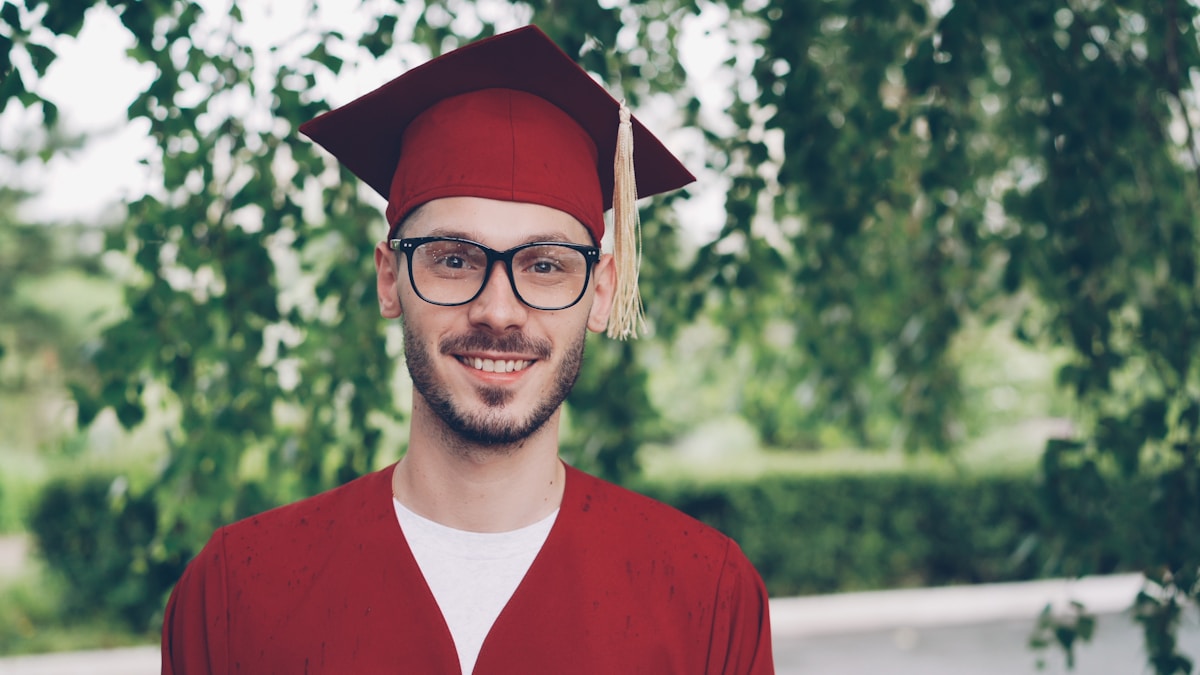 Homme diplômé souriant en robe de graduation rouge et lunettes.