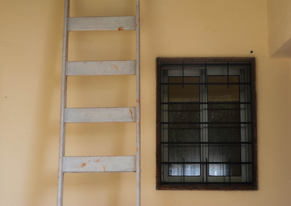 Échelle en bois, fenêtre grillagée, mur jaune.