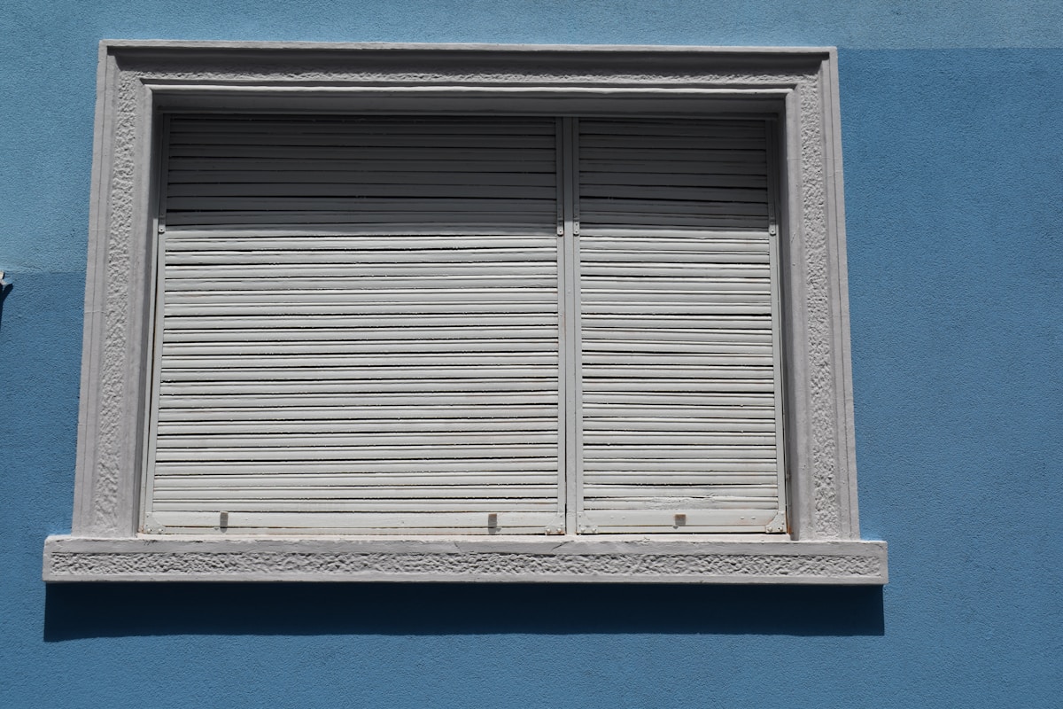 Fenêtre à volets métalliques gris sur mur bleu.
