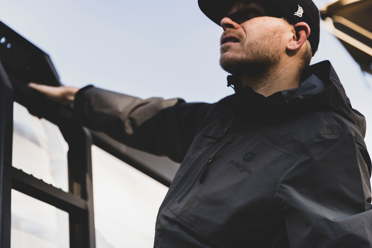Homme portant une veste noire avec logo, debout devant un paysage.