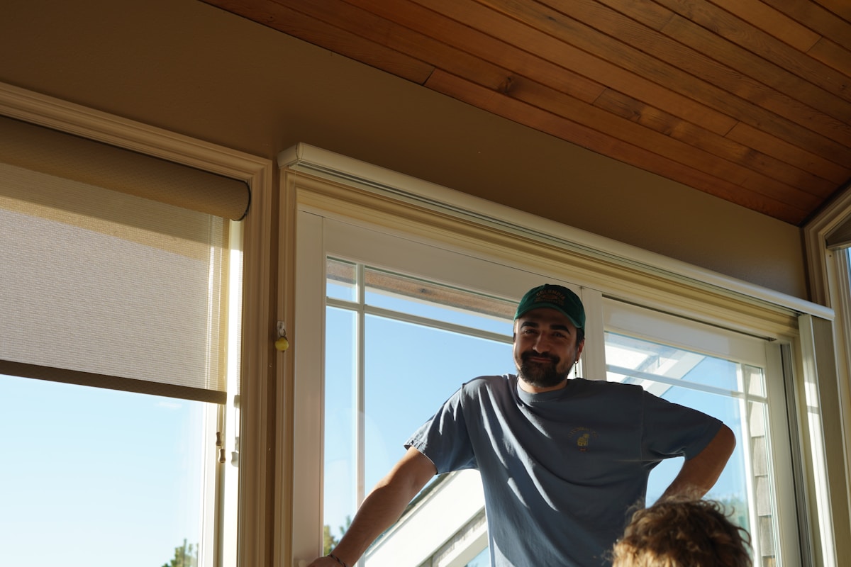 Homme barbu portant un chapeau, debout devant une fenêtre.