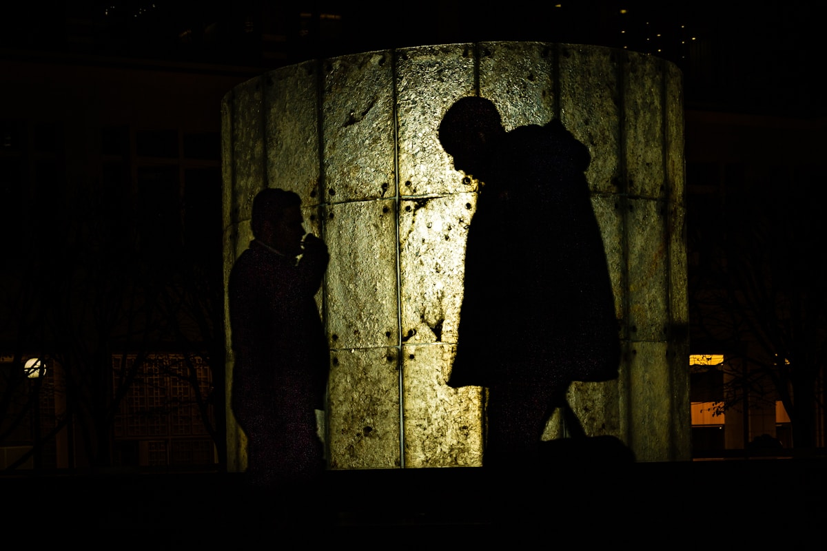 Deux silhouettes sombres se tiennent devant un mur de pierre éclairé.