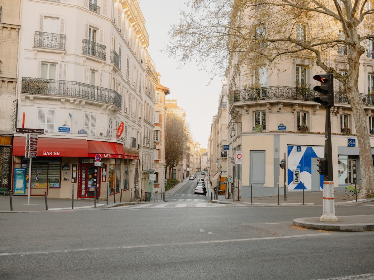 Rue parisienne avec bâtiments anciens, arbres, magasins et feux de circulation.