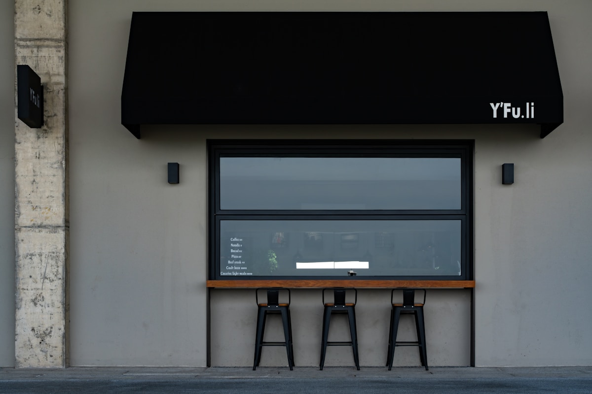 Façade minimaliste d'un établissement avec enseigne Y'Fu.li, fenêtre panoramique et tabourets noirs.