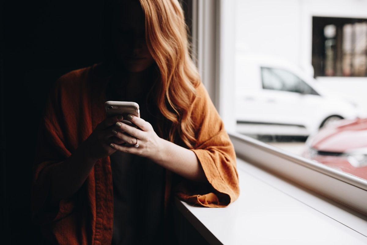 Femme aux cheveux roux utilisant un téléphone portable près d'une fenêtre.