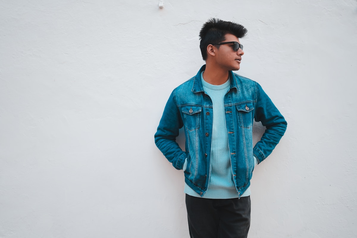 Homme portant une veste en jean bleu, debout devant un mur blanc.