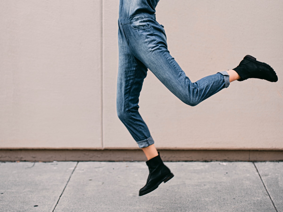 Jambes en mouvement, pantalon en jean, bottes noires, mur gris en arrière-plan.