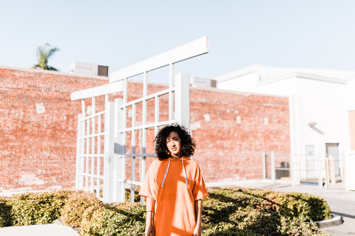 Femme aux cheveux bouclés portant un sweat orange devant un bâtiment en briques.
