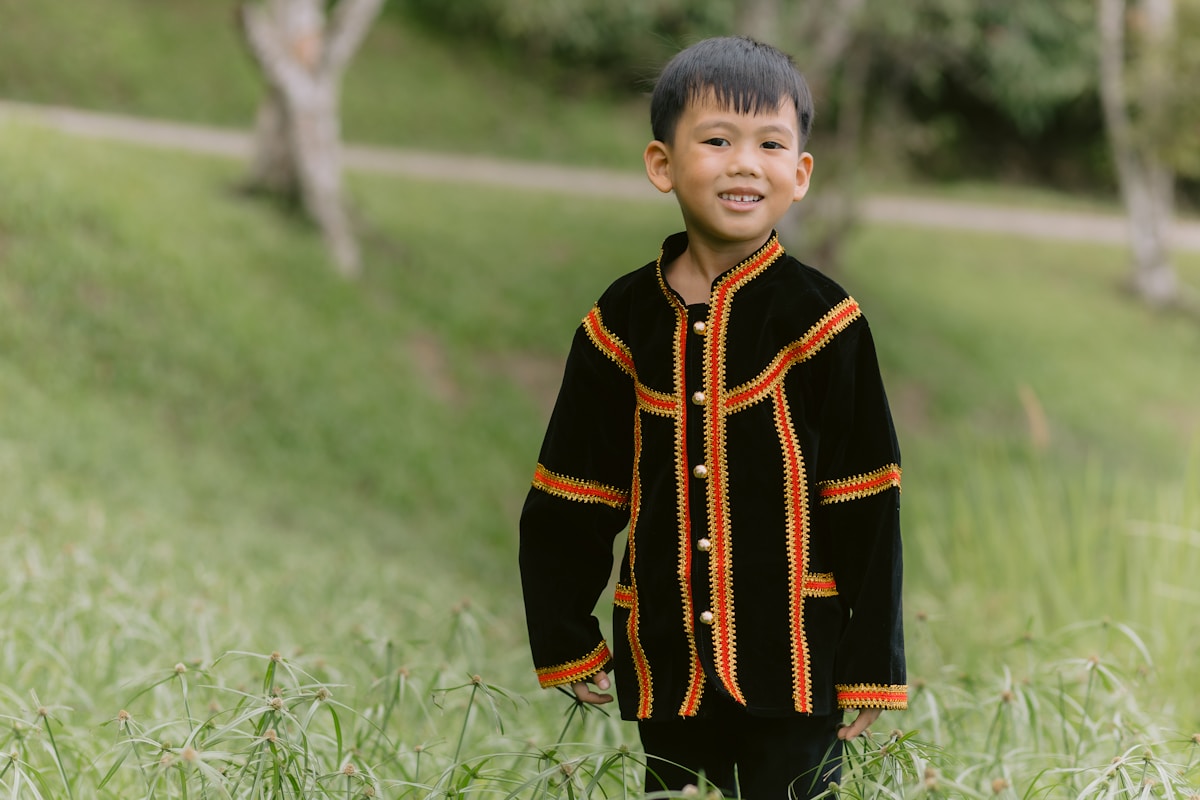 A smiling boy poses in traditional outfit outdoors.