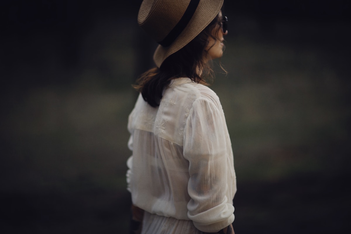 Femme en robe blanche et chapeau de paille, dans un environnement sombre.