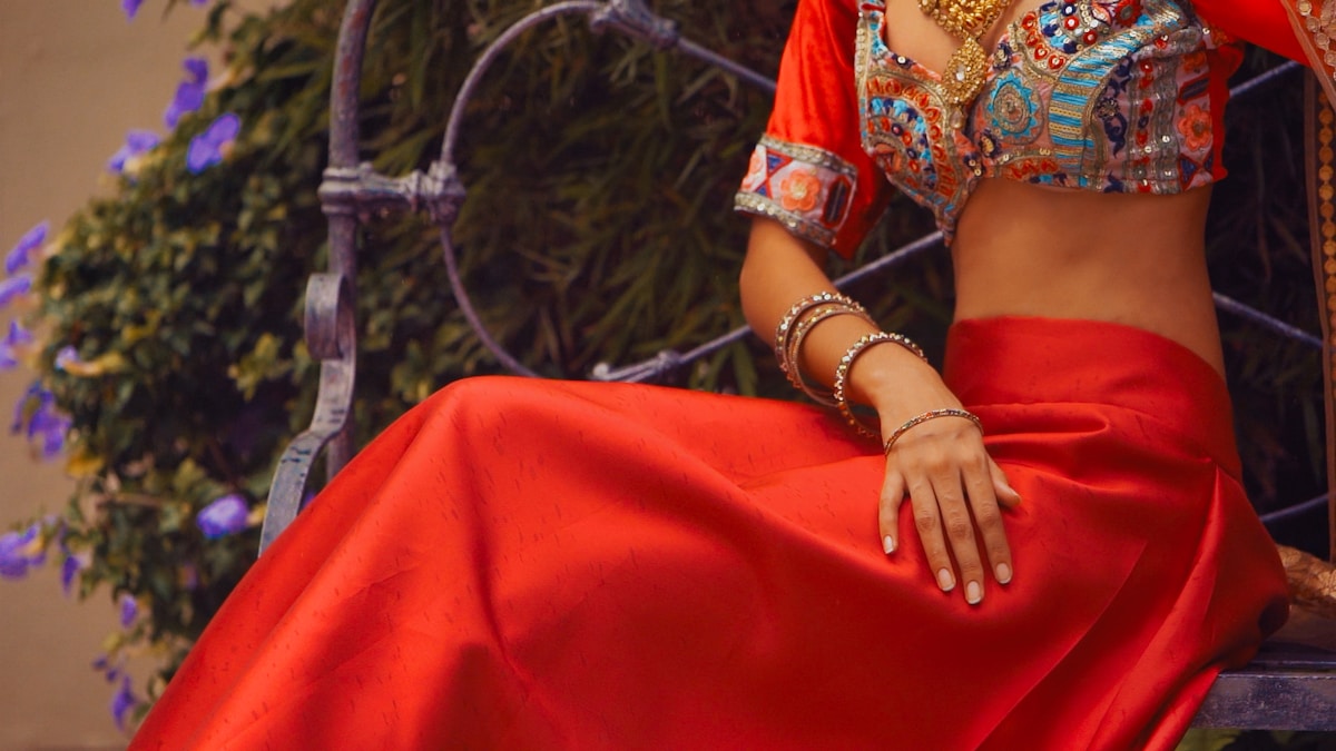 Femme en costume traditionnel rouge et doré, bijoux élégants, fond de verdure.