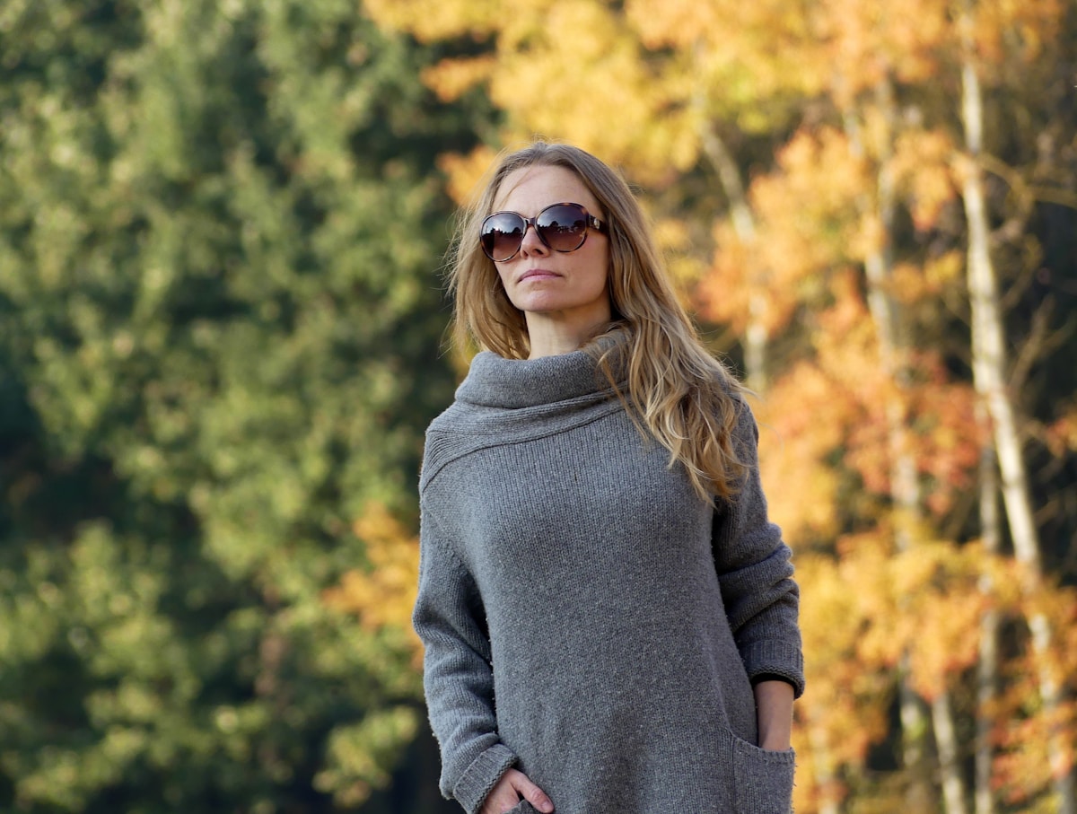 Femme aux cheveux blonds portant un pull gris dans un paysage automnal.