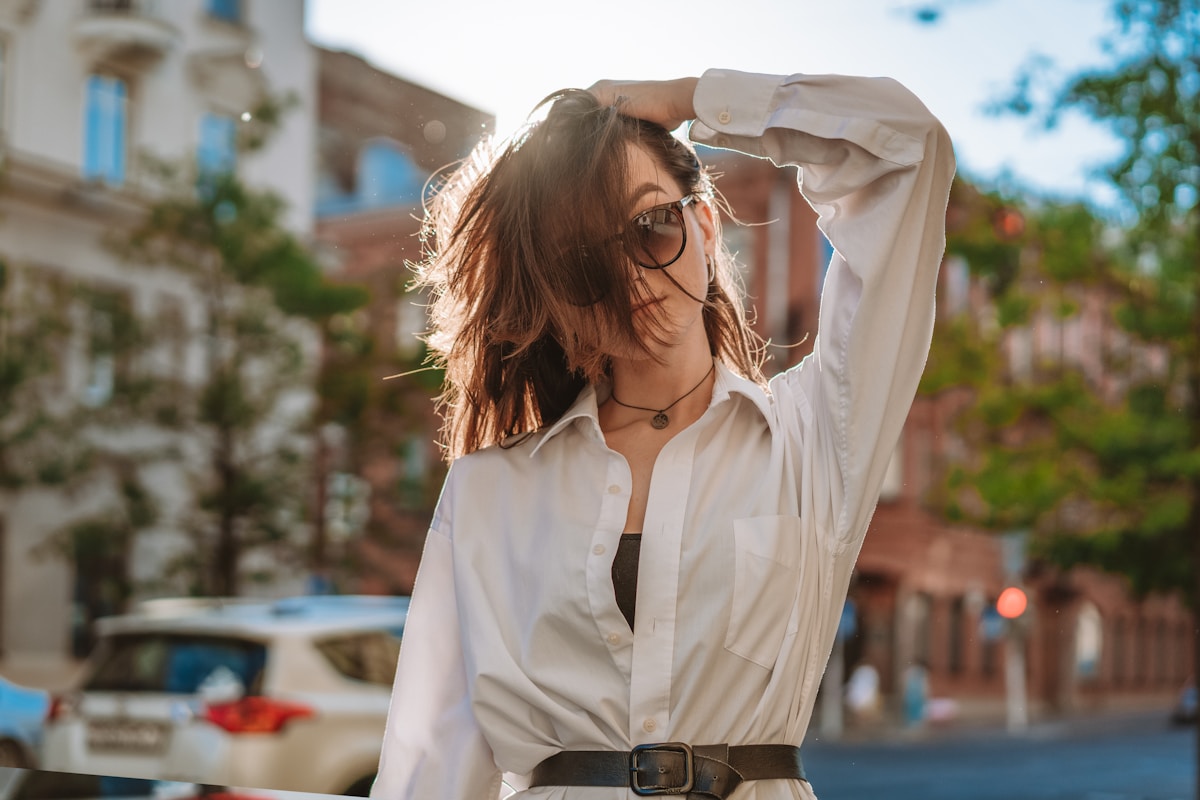 Femme aux cheveux bruns, lunettes et chemise blanche, dans une rue urbaine.