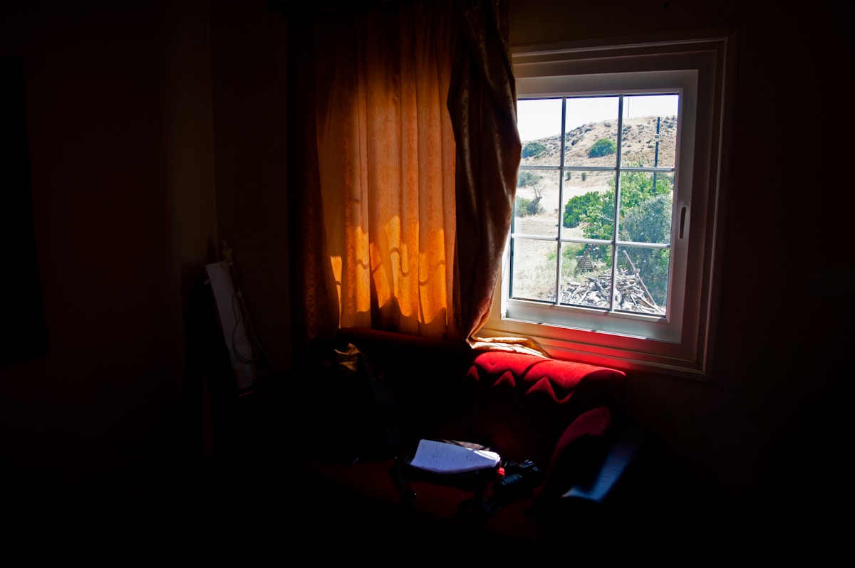 Fenêtre avec rideaux orangés, canapé rouge, paysage extérieur visible.