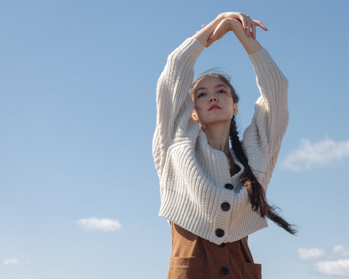 Jeune femme en pull blanc, bras levés, cheveux tressés, ciel bleu.