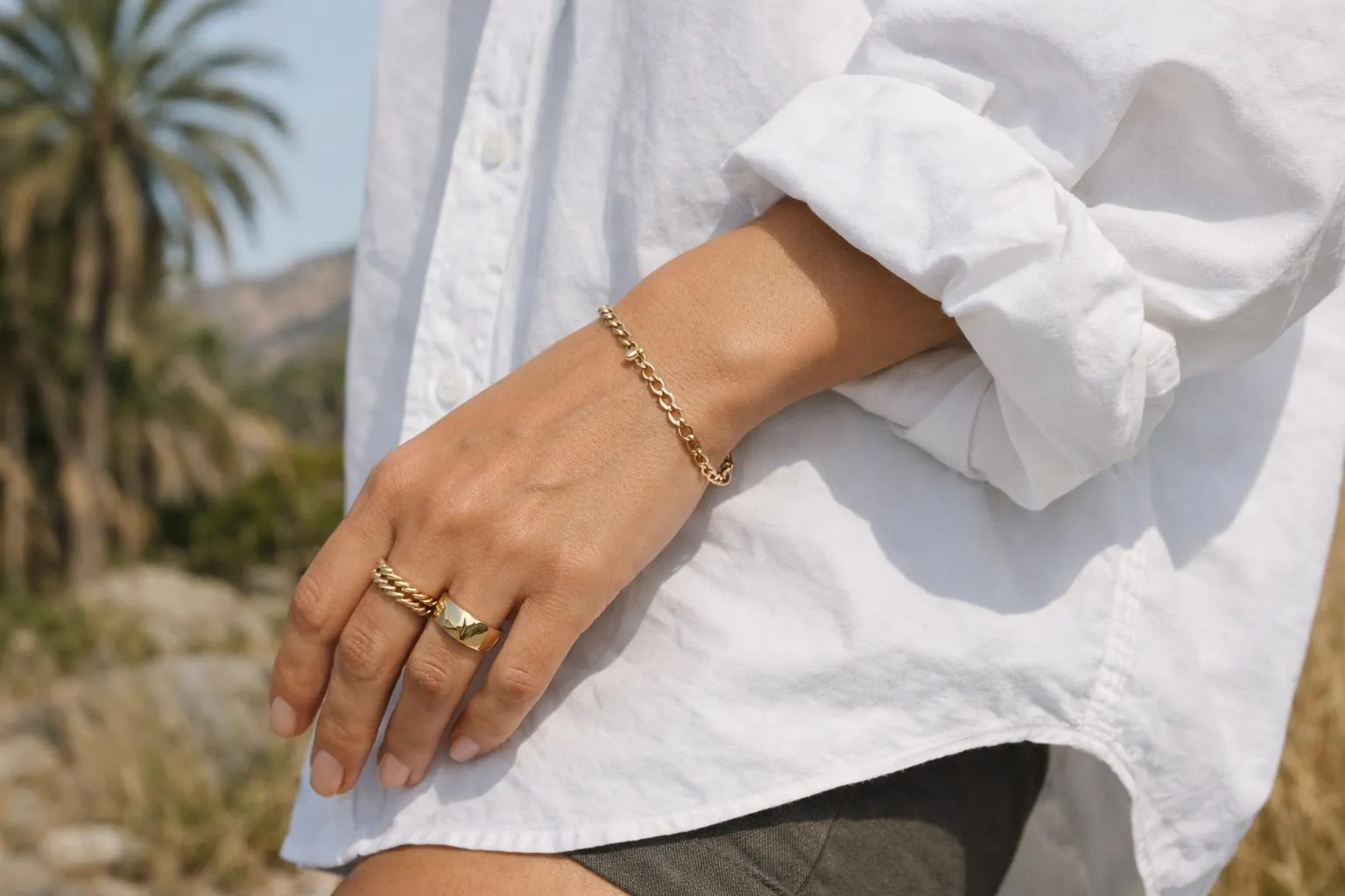 Femme portant une chemise blanche oversize décontractée, manches retroussées, combinée avec un jean déchiré et des accessoires bohèmes dorés, dans un décor lumineux californien avec végétation