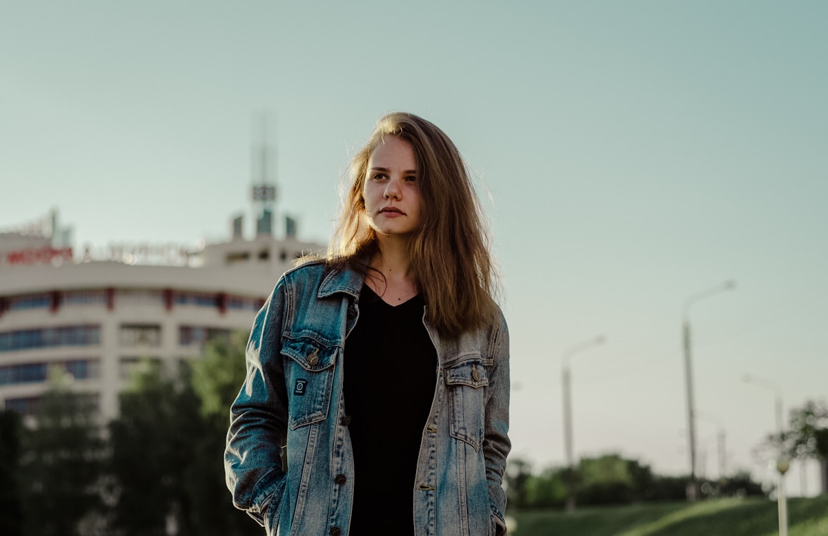 Jeune femme aux cheveux longs portant une veste en jean, debout devant un paysage urbain.