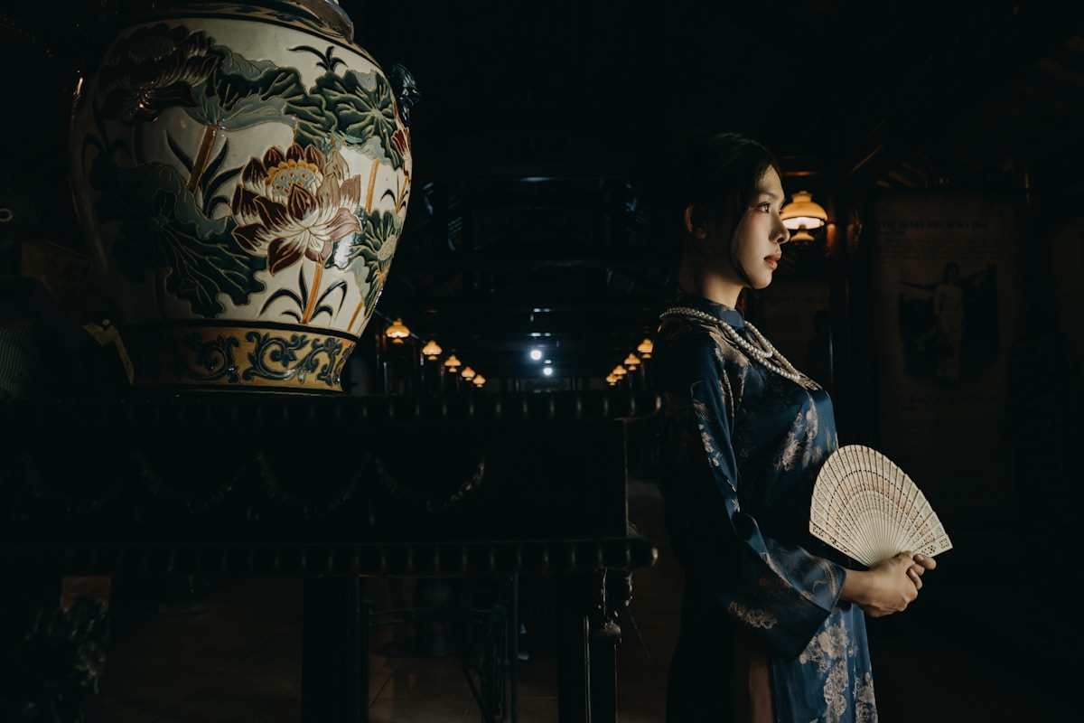 Femme en kimono bleu tenant un éventail, devant un vase décoré de fleurs.
