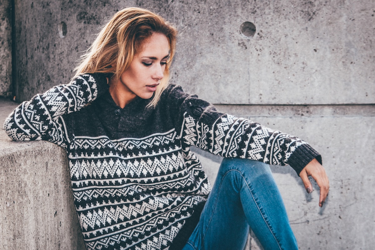 Femme blonde pensive, vêtue d'un pull à motifs nordiques, assise contre un mur gris.
