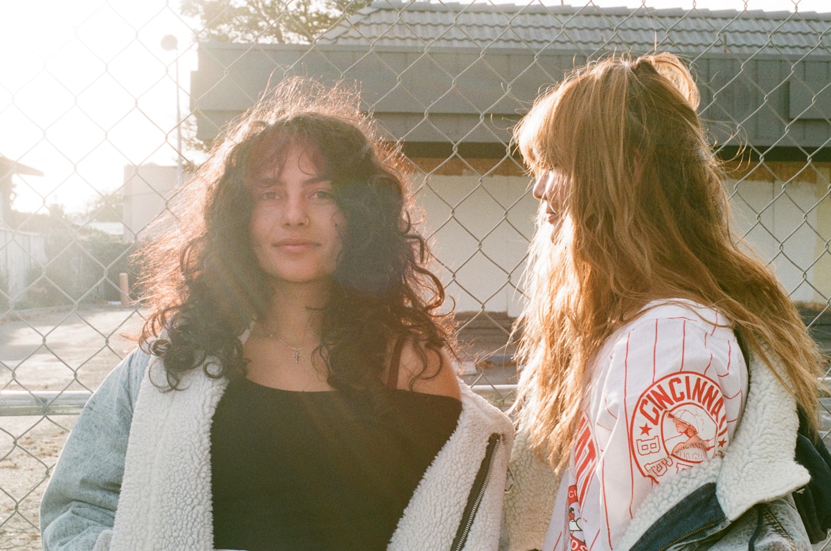 Deux jeunes femmes aux cheveux longs, l'une brune et l'autre blonde, posant devant une clôture.