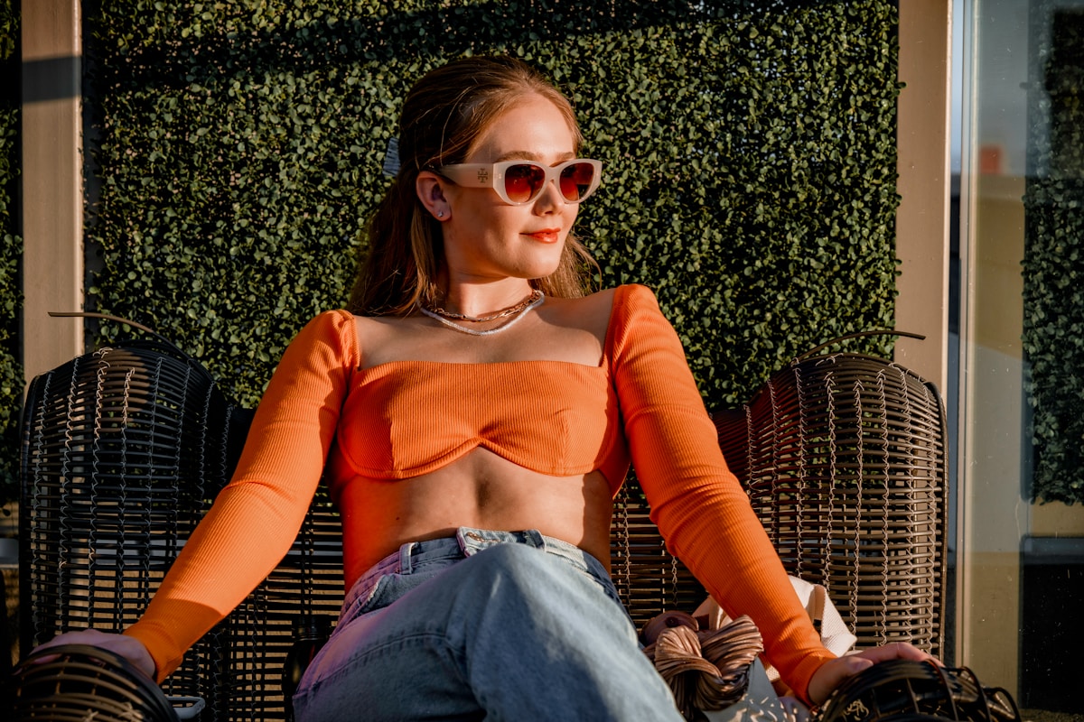 Femme aux cheveux bruns portant des lunettes de soleil rouges et un haut orange.