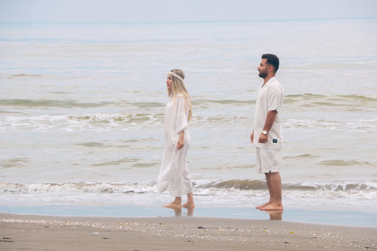 Couple en tenue blanche marchant sur la plage, mer calme en arrière-plan.
