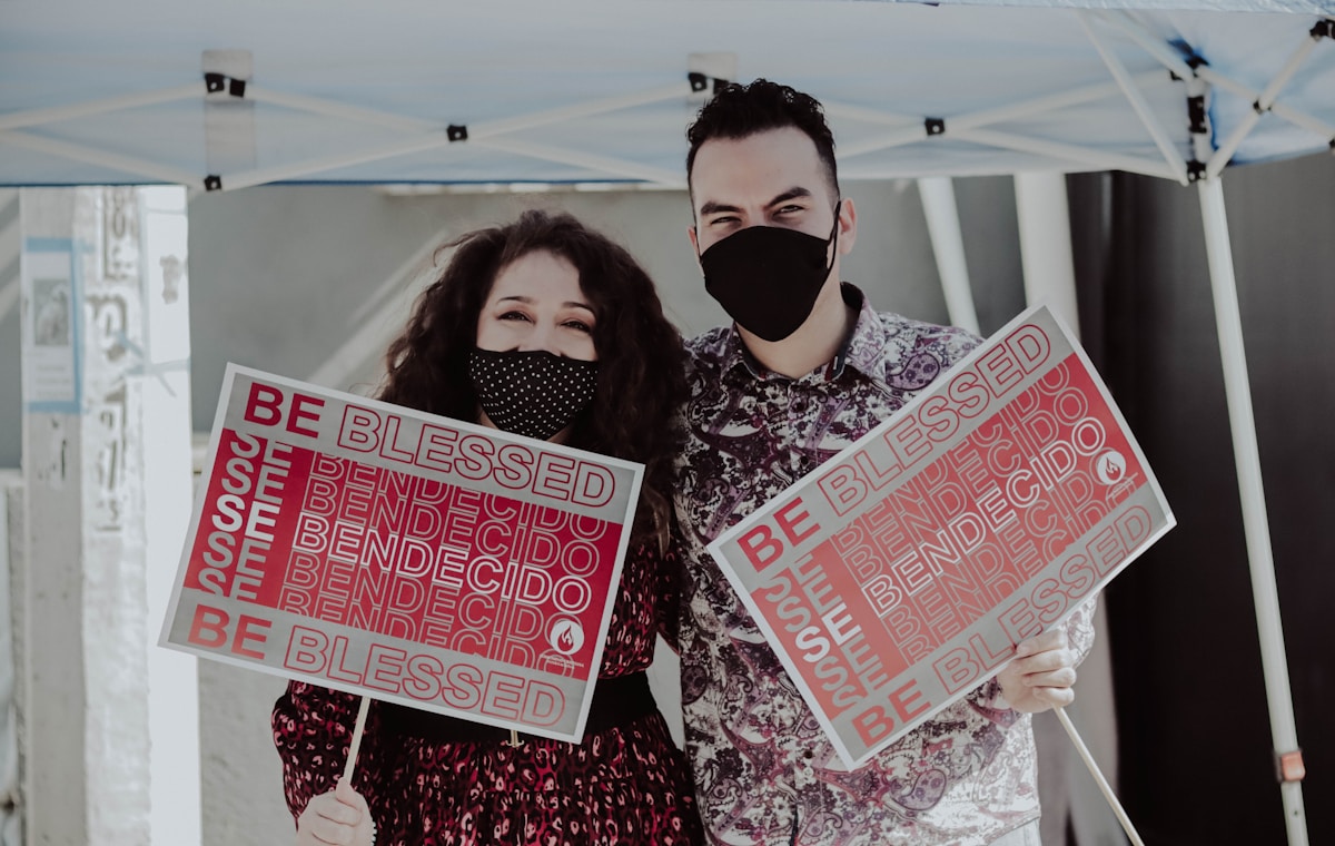 Deux personnes masquées tenant des pancartes "Be blessed" dans un environnement extérieur.
