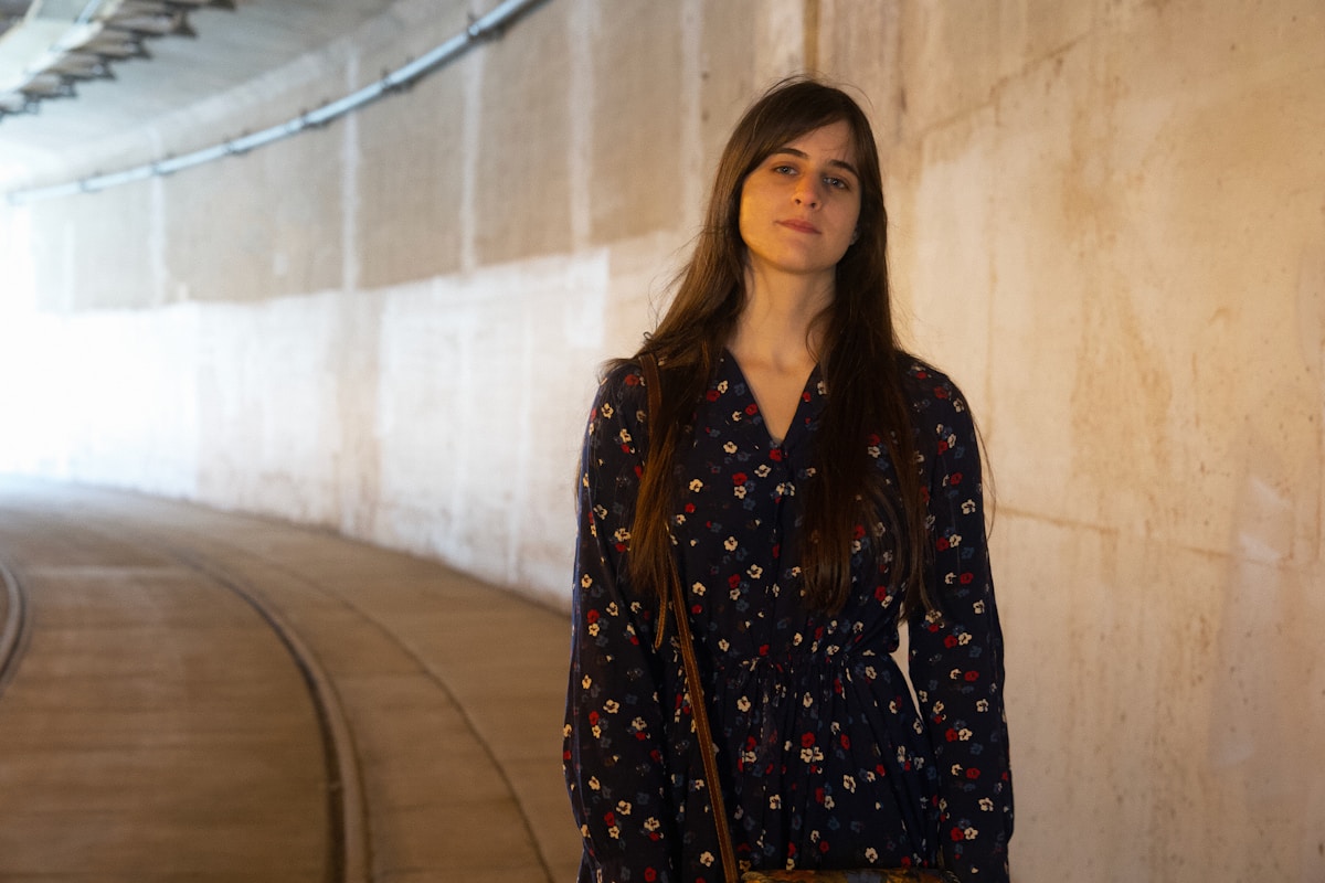 Jeune femme aux longs cheveux bruns portant une robe fleurie dans un couloir sombre.